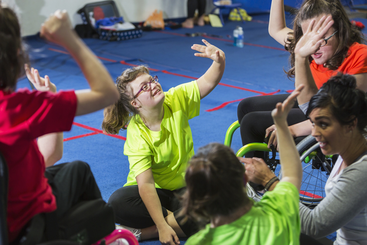 Teenage girl sitting on a gym floor with a group of other girls. She is smiling, wearing a bright yellow shirt, raising her hand.