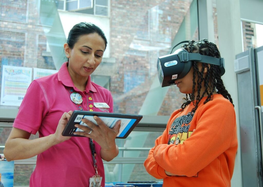 Play Specialist Shirin in pink Evelina t-shirt on the left holding an tablet to control the VR game, Oliver is in an orange hoodie wearing the VR head set.