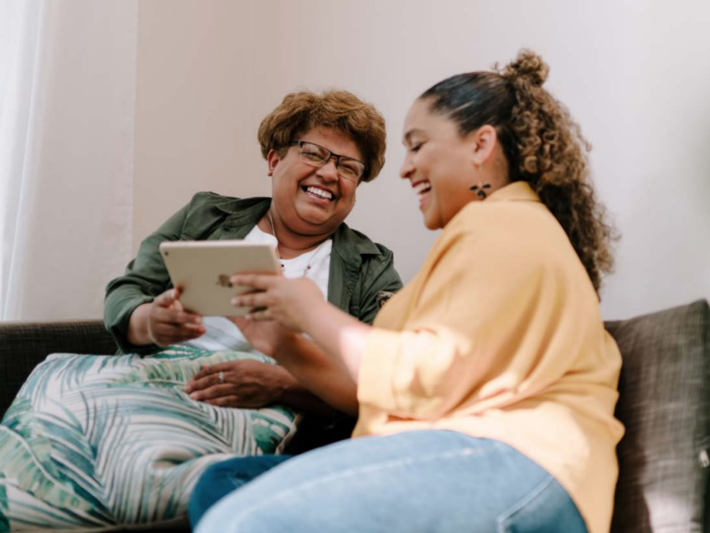 Two women holding an ipad, they are sat on the sofa and appear to be smiling at each other.
