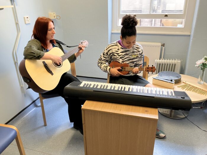 Kiefer on the right sat down wearing a dark brown and grey tiger print jumper looking down smiling whilst playing a dark brown ukulele. On the left is Hannah also sat down wearing a dark green shirt, holding a light brown guitar.