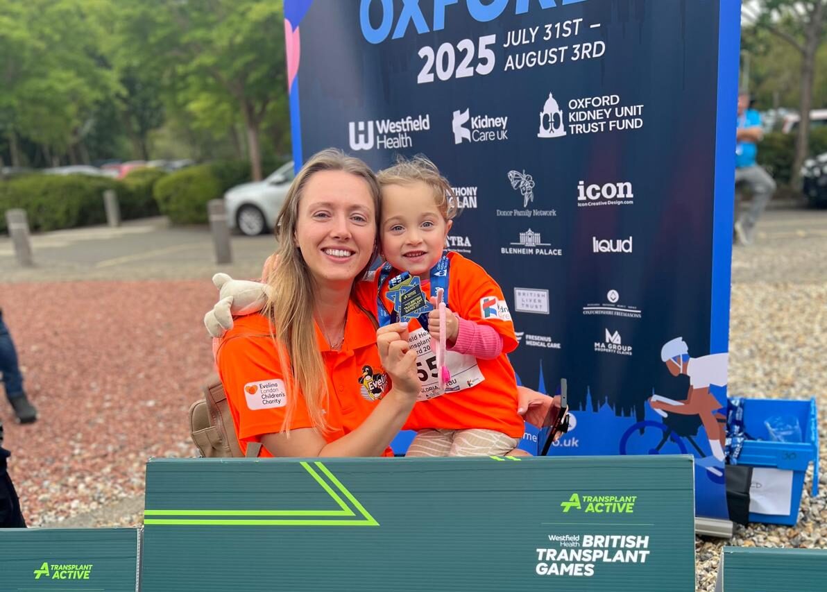 Mum, Laura holding her little girl Gloria who is holding up her medal. Both are in bright orange shirts and on Laura's shoulder there is a badge that reads 'Evelina London Children's Charity'. They are behind a green board that reads 'British Transplant Games'