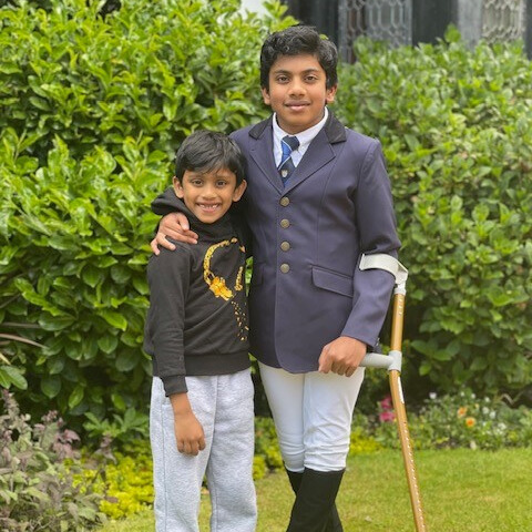 Govind on the right in a navy blazer with a crutch with his arm round his younger brother, both are in front of a green hedge