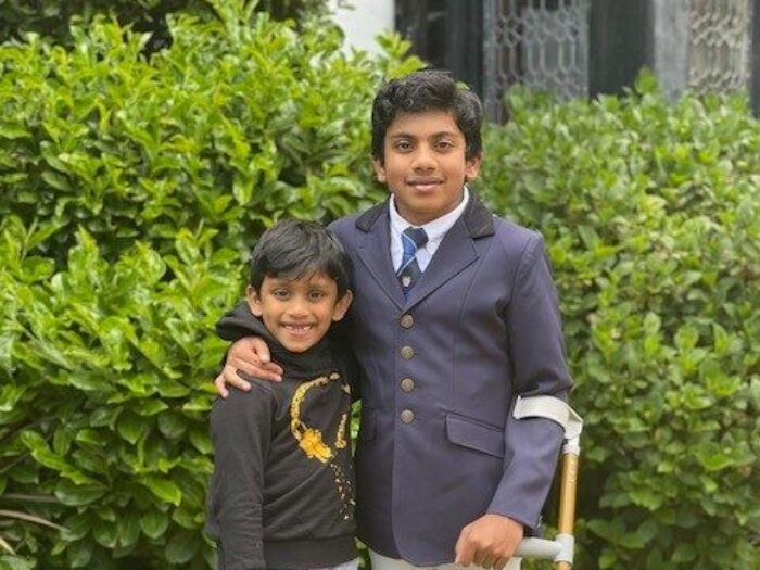 Govind on the right in a navy blazer with a crutch with his arm round his younger brother, both are in front of a green hedge