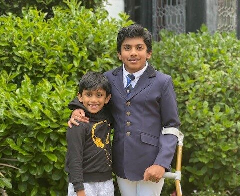 Govind on the right in a navy blazer with a crutch with his arm round his younger brother, both are in front of a green hedge