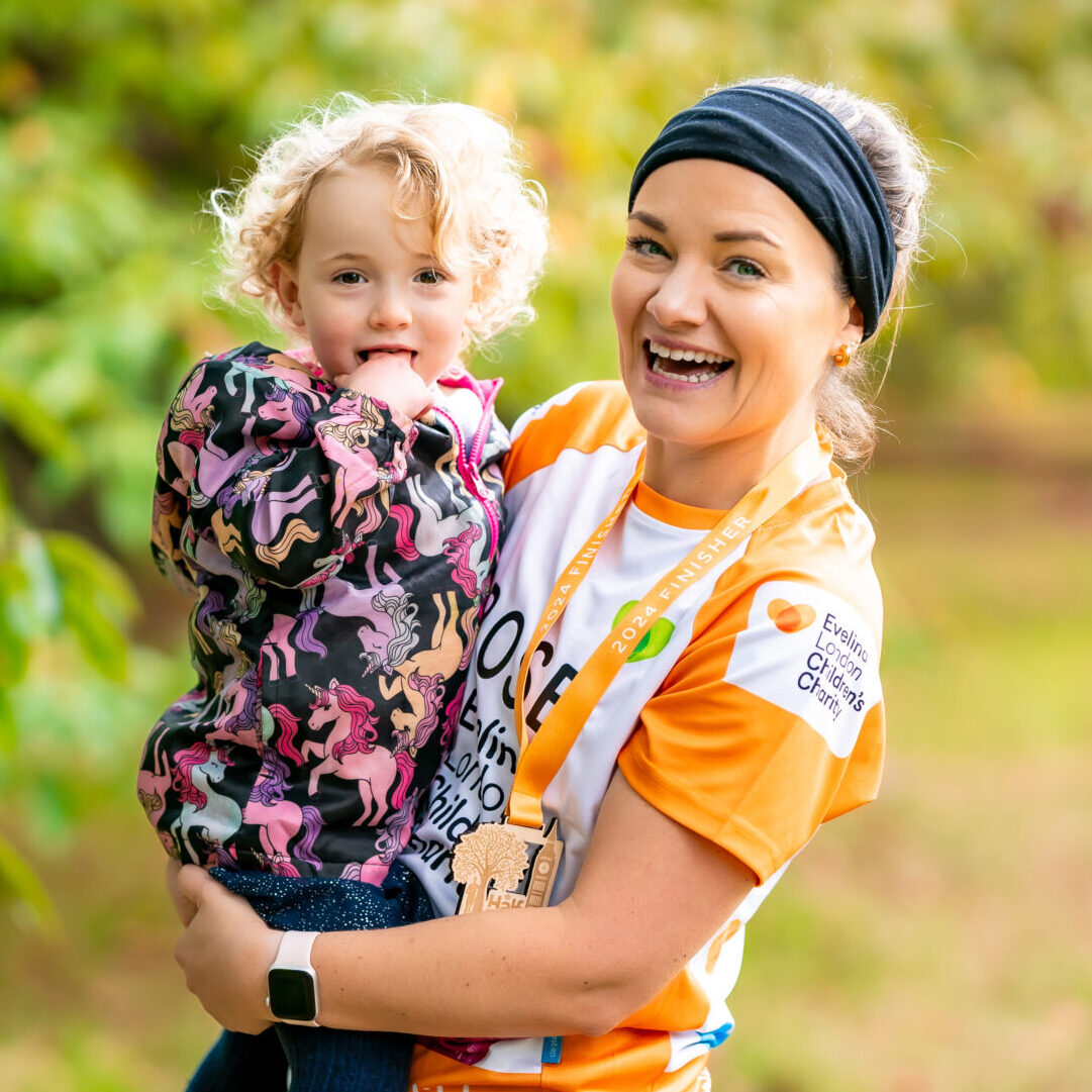 Rose smiling wearing a black headband and an orange Evelina London Children's Charity T-Shirt, holding her daughter Molly who is wearing a jacket with ponies on it. Both are in front of blurred bush background.