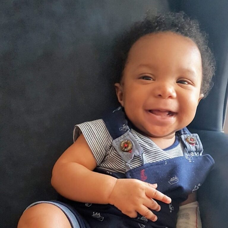 Baby Hamza smiling in a striped t shirt with navy dungarees on top with boats printed on them.