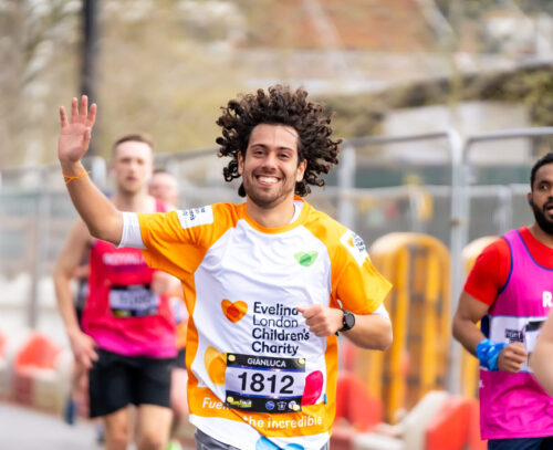 Man smiling and giving a wave whilst running a marathon wearing an Evelina London Children