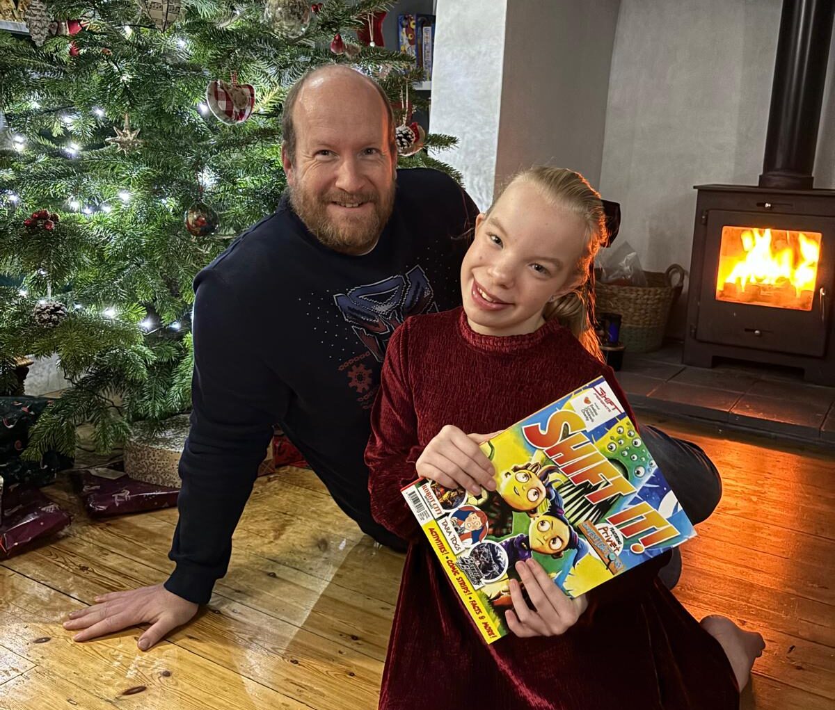 Naia with her dad, Adrian holding the comic book with Christmas tree and fireplace behind them.