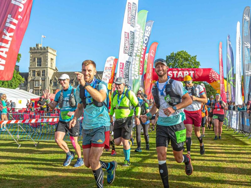 Ultra challenges at the start just setting off on their run, walk or jog. They're all wearing running vests, in the background there is numerous flags of different charities.