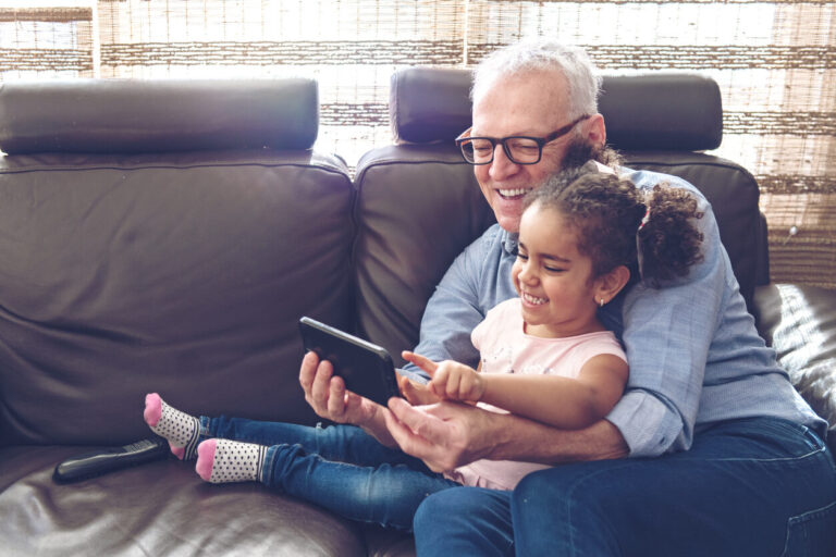 Grandfather playing with granddaughter sat on the sofa watching a mobile