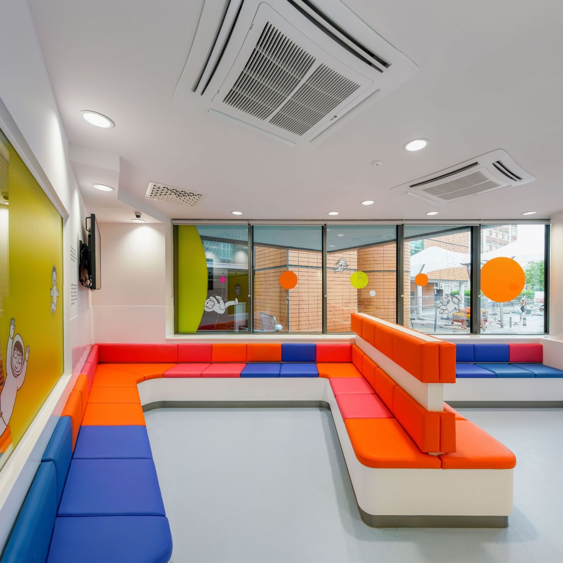 Evelina London Children's hospital waiting room with colourful orange, red and blue chairs. On the walls and windows feature black and white astronaut illustrated stickers and orange circles.