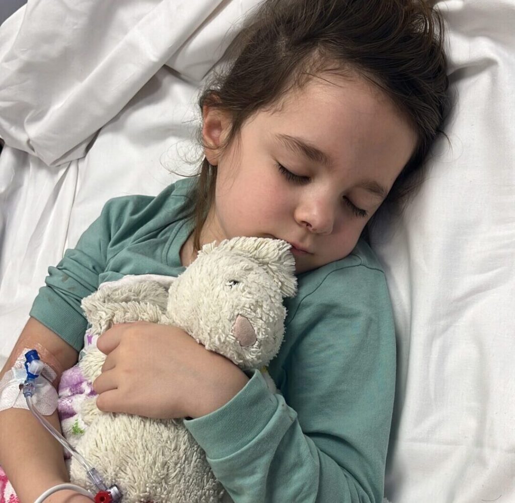 Indy cuddling a soft cream plush wearing a green jumper asleep in Evelina London Hospital bed.