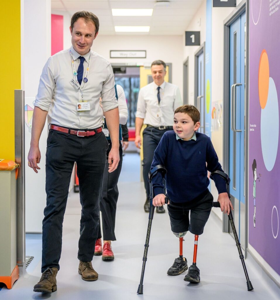Tony Hudgell on the right wearing a blue jumper with crutches walking next to a member of staff from Evelina London Day Centre on the left who is wearing a white shirt with rolled up sleeves with a blue tie, lanyard with his staff badge around his neck and what appears to be dark blue trousers, there is also two members of staff walking behind them.