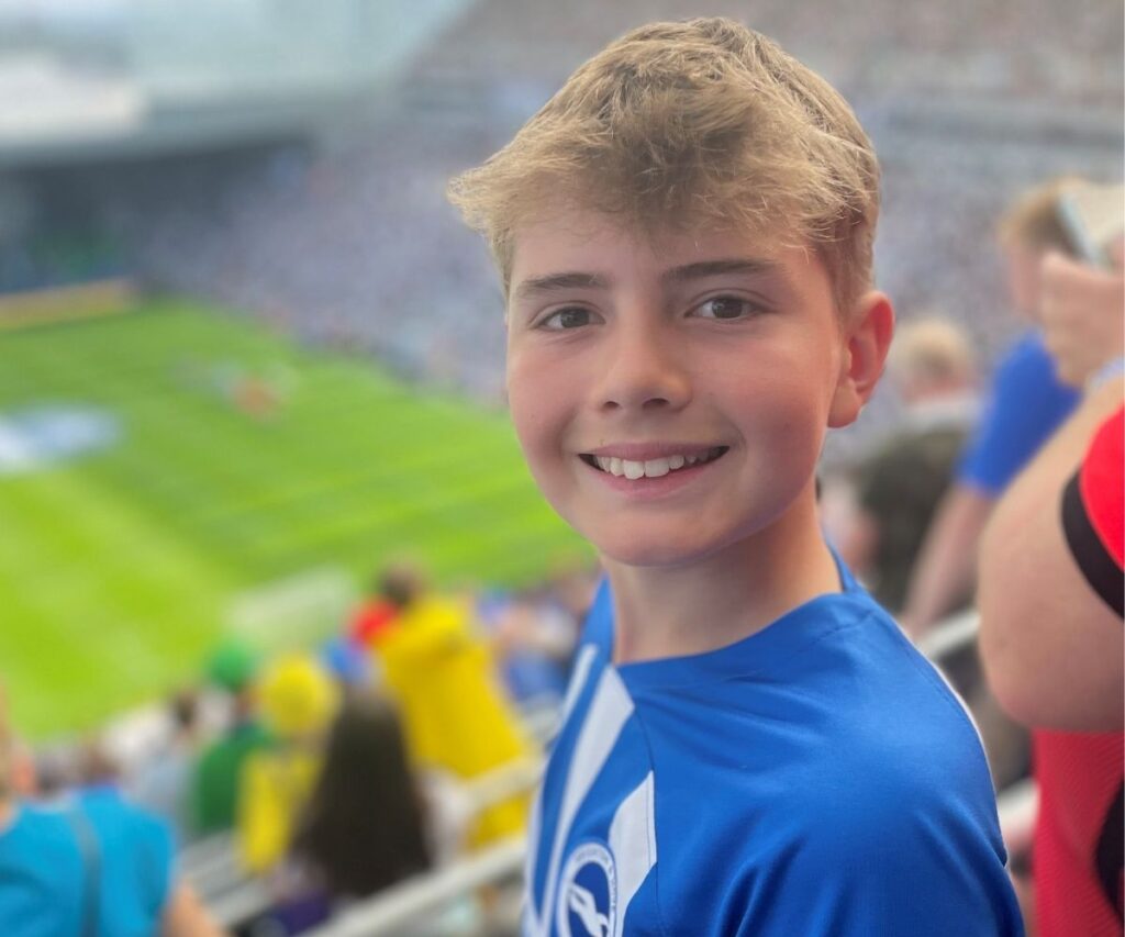 Austin smiling wearing a blue and white striped Brighton and Hoves football t-shirt amongst the crowd waiting to see the football match.