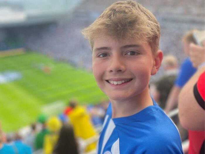 Austin smiling wearing a blue and white striped Brighton and Hoves football t-shirt amongst the crowd waiting to see the football match.