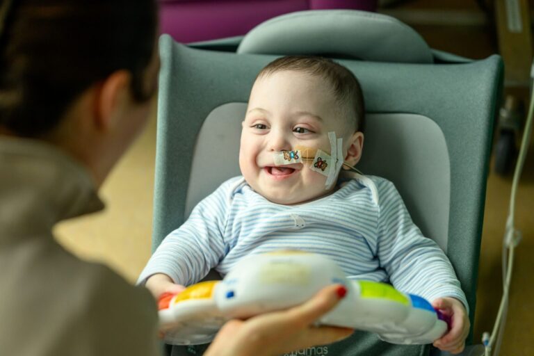 Bertie smiling sat on a chair, looking at mum Phoebe who is on the left holding up a colourful toy.