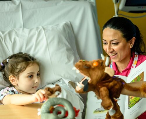 Evelina Play Specialist Shirin holding a Gruffalo hand puppet with the Gruffalo story book telling Olilia the story while shes sat up in hospital bed at Evelina London Children