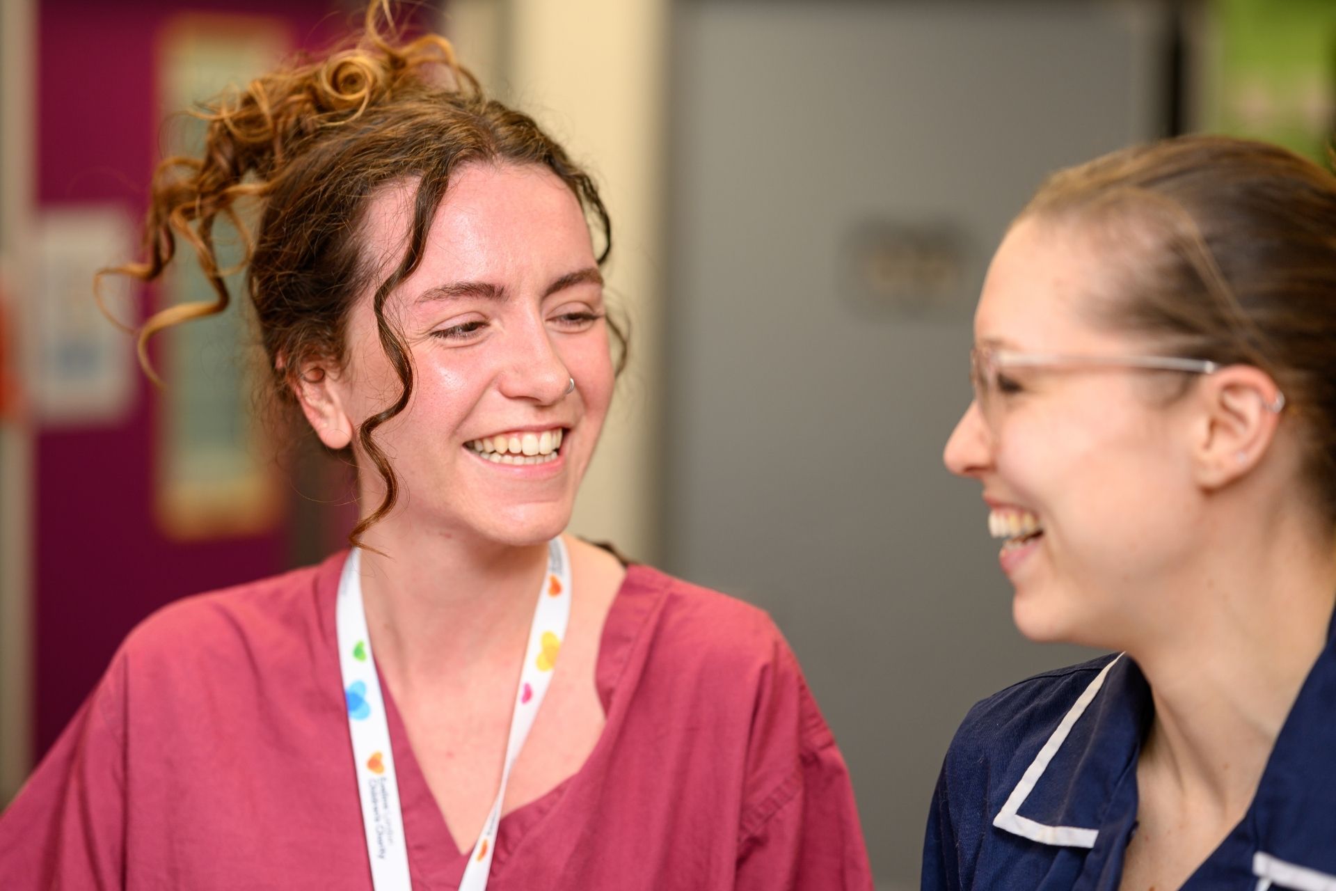 Two members of Evelina London Children's Hospital staff walking and smiling and chatting to each other. On the left she's wearing a pink uniform with a white Evelina London Charity's lanyard and on the right she's wearing light brown glasses, wearing a blue uniform.