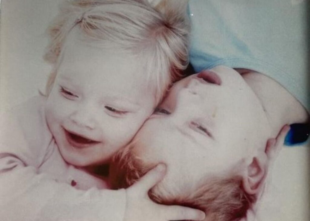Jasmine and Jordan lying down next to eachother, Jasmine on the left in pink holding her brother Jordans head appearing to be smiling.