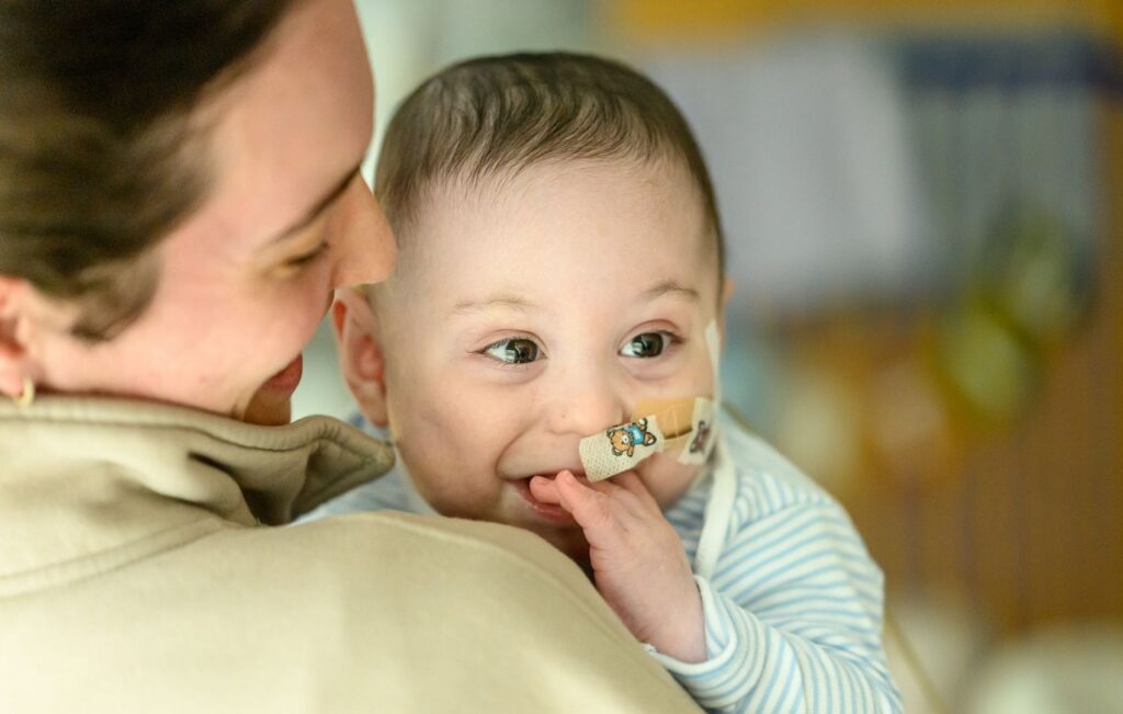 Baby Bertie being carried by his mum, Bertie has his fingers in his mouth and appears to be smiling.