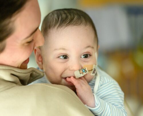 Baby Bertie being carried by his mum, Bertie has his fingers in his mouth and appears to be smiling.