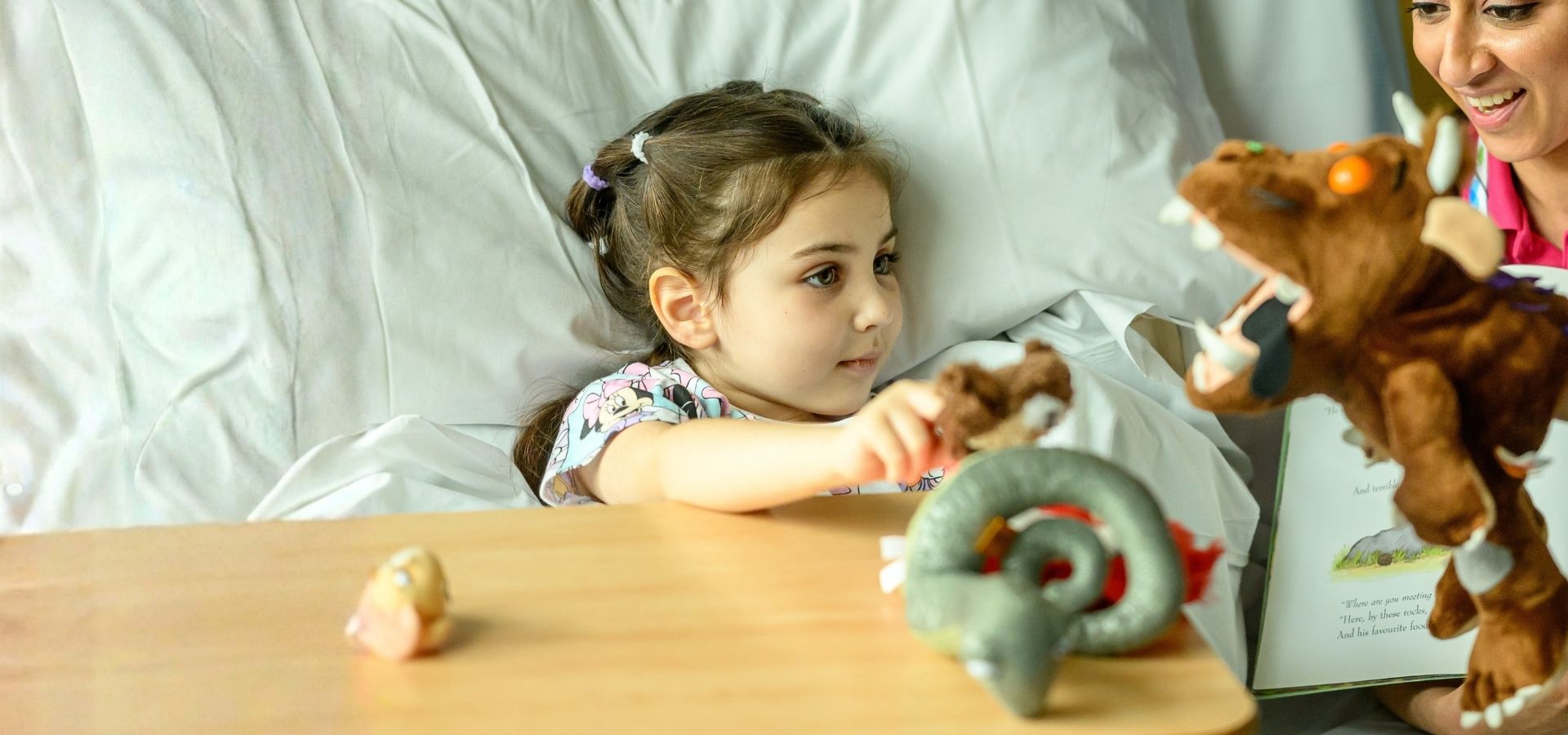 Olilia in the hospital bed at Evelina London Children's Hospital with Evelina play specialist, Shirin. Shirin is telling her about the story of the Gruffalo with a brown Gruffalo hand puppet.