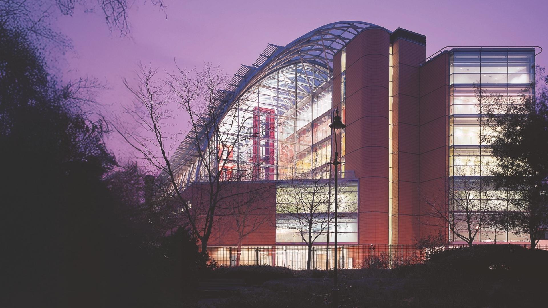 Evelina London Children's Hospital mainly made of glass with lights on at night with trees on the left side.