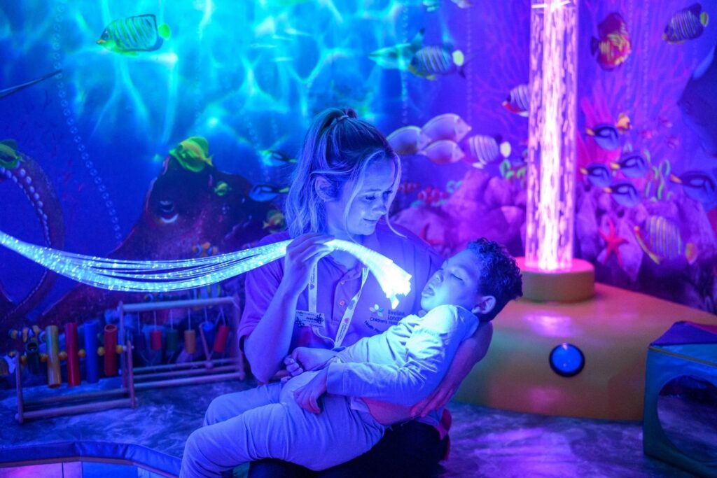 In the sensory room, Jess an Evelina London Play Specialist is holding strands of lights, in her arms she