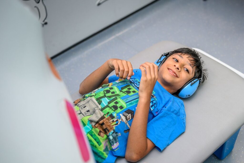 A boy lying down in an MRI scanner who appears to be smiling, he is wearing a blue head set and wearing a blue t shirt with green blocks with faces on them.