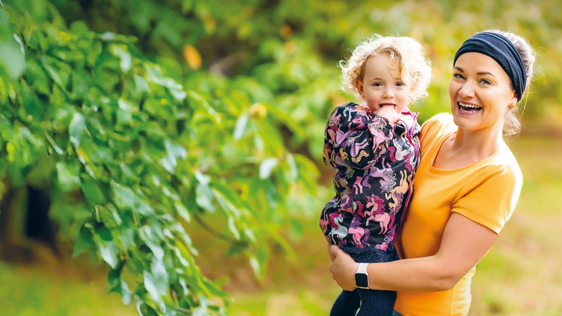 A mother smiling wearing a black headband and an orange T-Shirt, holding her daughter who is wearing a jacket with ponies on it. Both are in front of blurred bush background.
