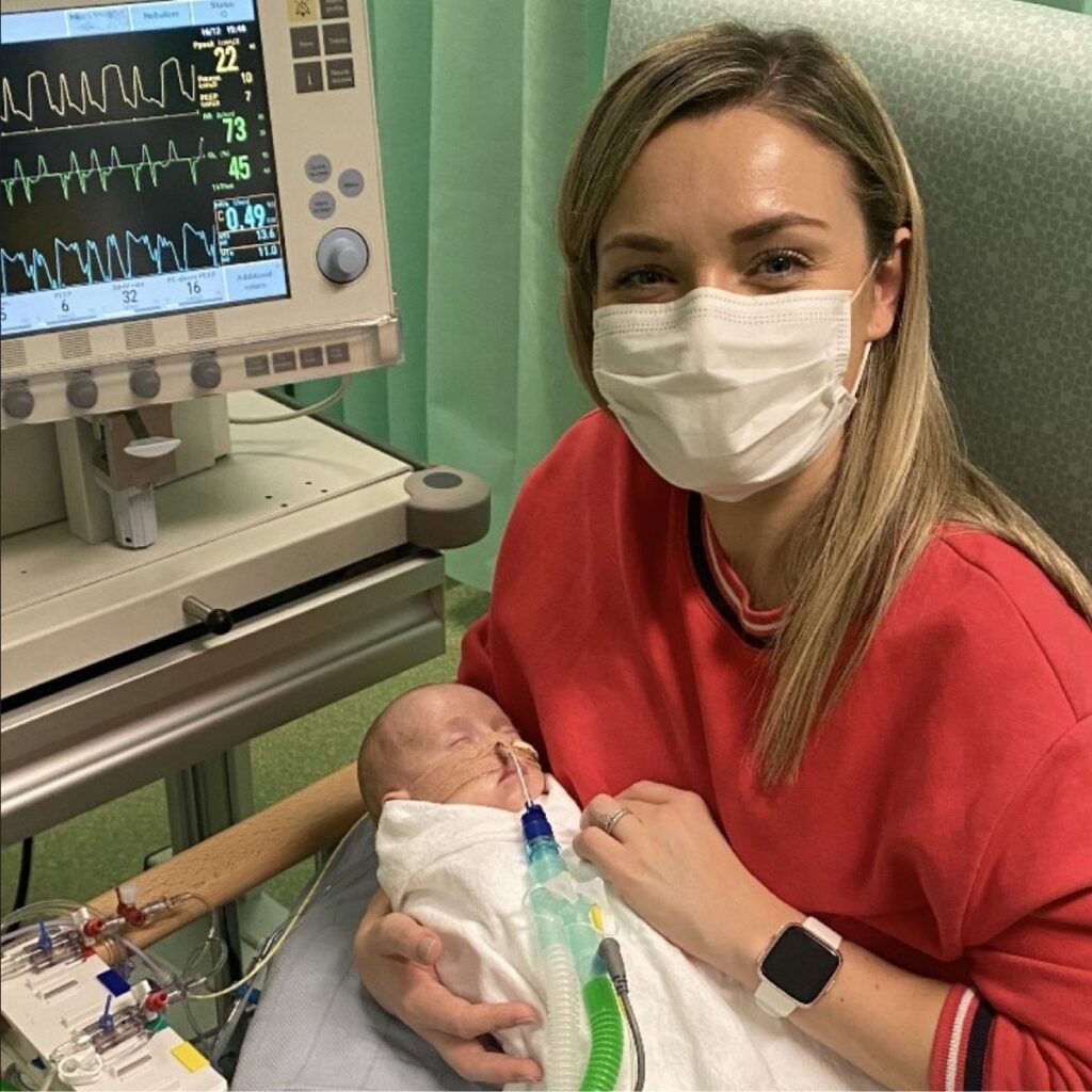 Rose wearing a white facemask and red jumper, holding a baby in her arms who is connected to a hospital machine.