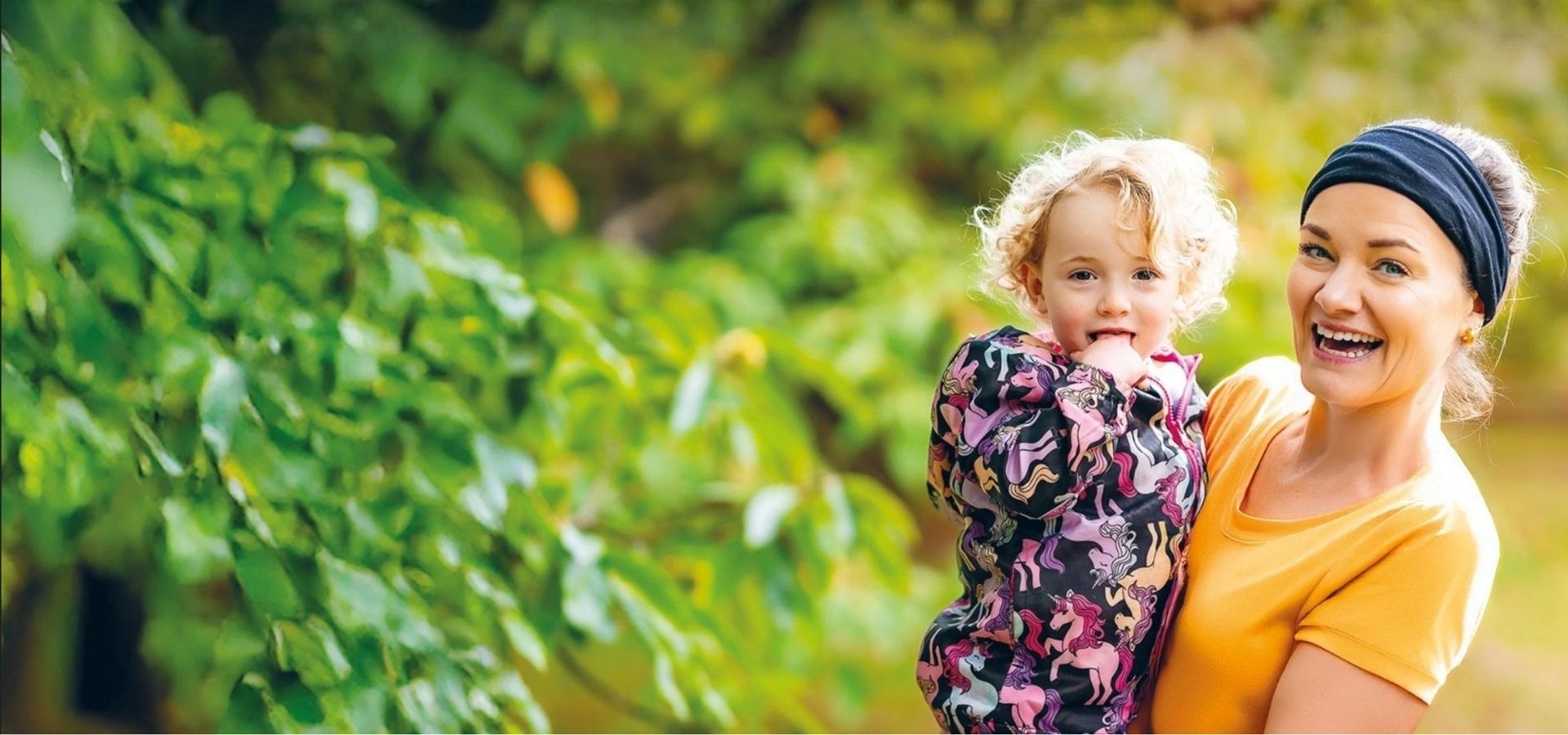 A mother smiling wearing a black headband and an orange T-Shirt, holding her daughter who is wearing a jacket with ponies on it. Both are in front of blurred bush background.