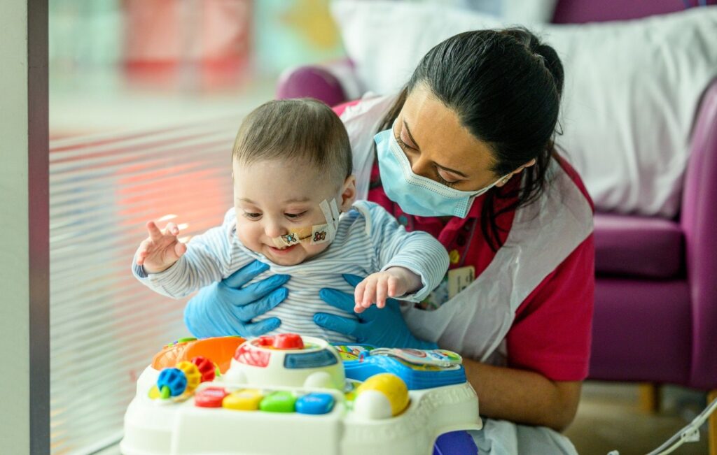Baby Bertie being held by Play Specialist Shirin, he is playing with a colourful development toy which has colourful buttons.