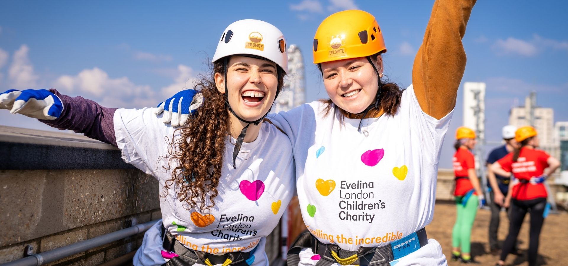 Two women with their arms up in joy wearing helmets and white t-shirt that reads 'Evelina Children's Charity'