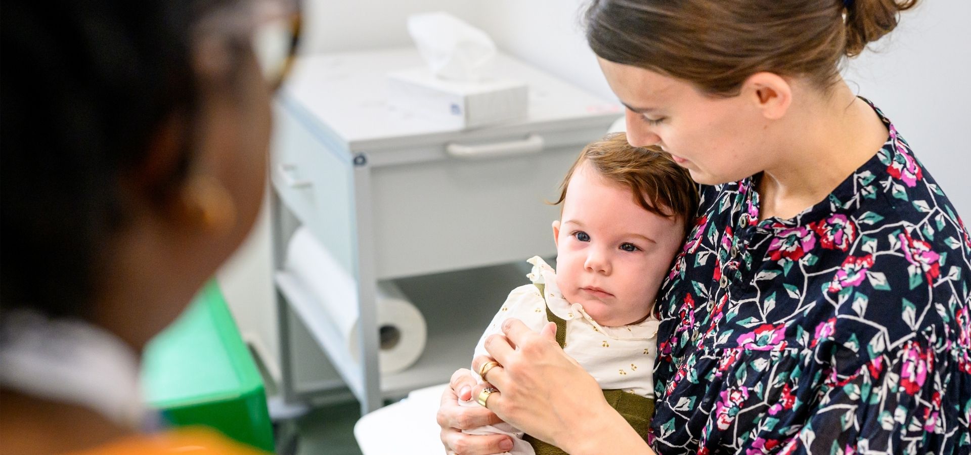 Mother holding her baby at an appointment in Evelina London Children's Hospital. Mum is wearing a floral print top and baby on her lap is in a white floral top with green overalls.