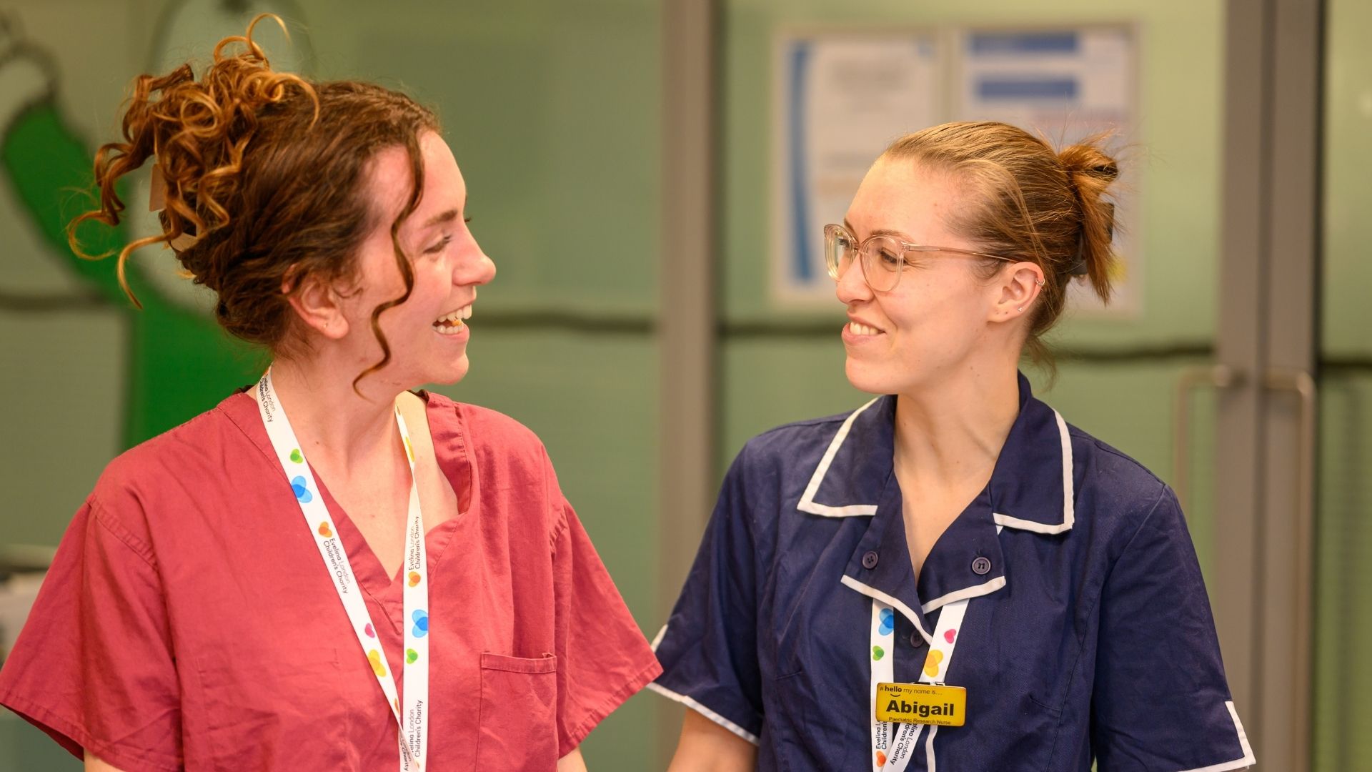 Two members of Evelina London Children's Hospital staff walking and smiling and chatting to each other. On the right she's wearing a blue uniform with a yellow name badge on her lanyard that say's 'Abigail' and on the left she's in a pink uniform.