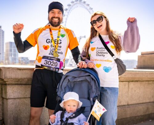 A family of three Evelina London Charity Supporters, the two parents have their arms up in the air cheering, their child is in their pram. Behind them is blue sky and The London Eye.