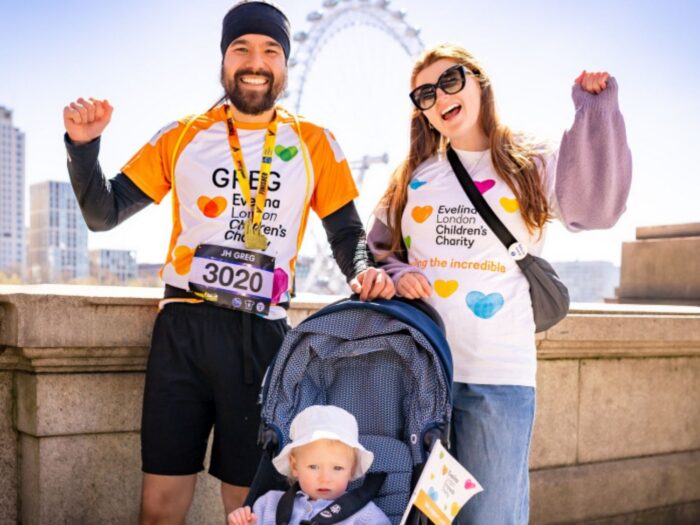 A family of three Evelina London Charity Supporters, the two parents have their arms up in the air cheering, their child is in their pram. Behind them is blue sky and The London Eye.