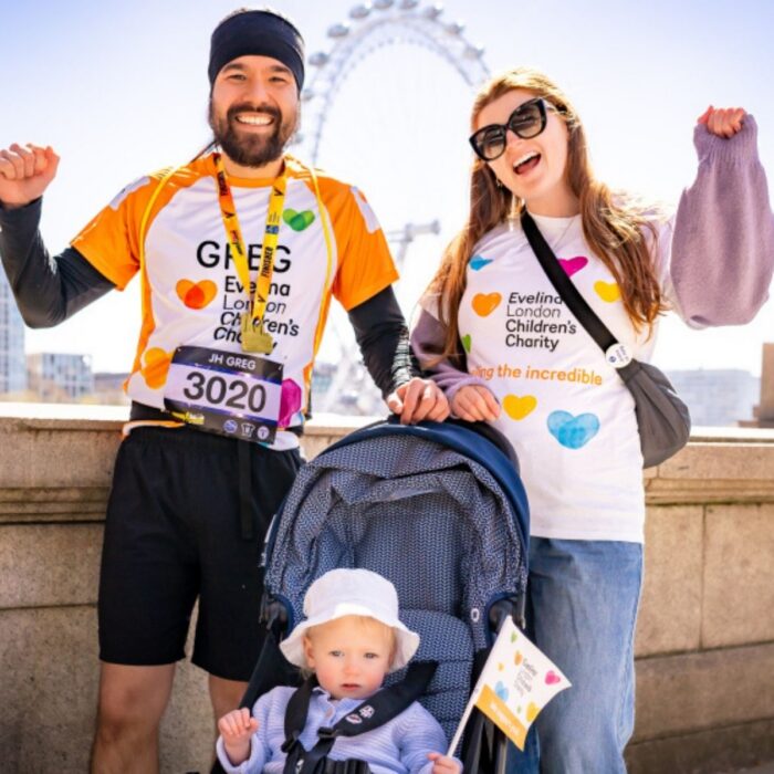 A family of three Evelina London Charity Supporters, the two parents have their arms up in the air cheering, their child is in their pram. Behind them is blue sky and The London Eye.