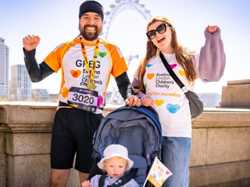 A family of three Evelina London Charity Supporters, the two parents have their arms up in the air cheering, their child is in their pram. Behind them is blue sky and The London Eye.