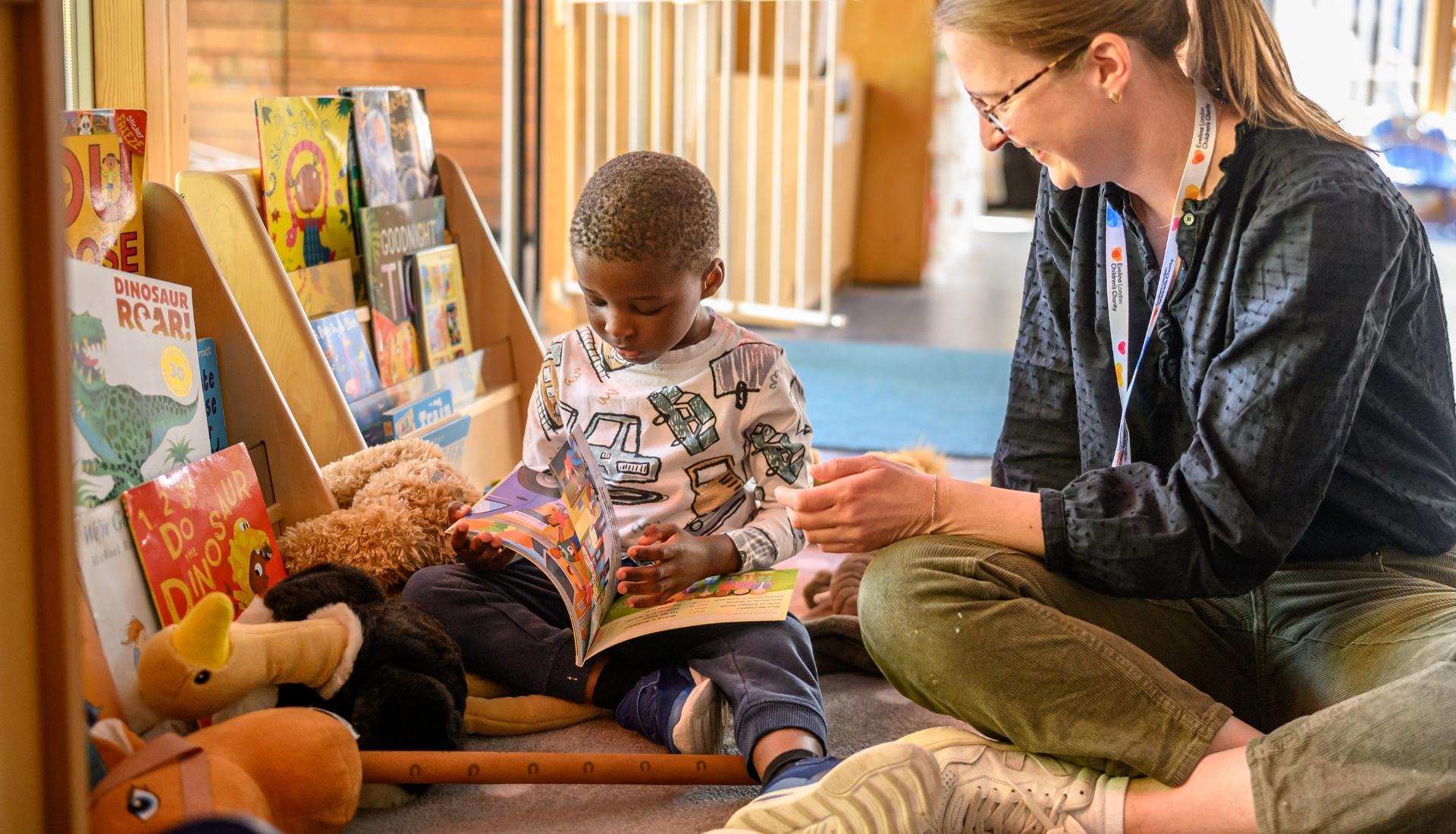Child sat on the floor amongst lots of storybooks, he's reading a story and besides him is an Evelina London Children's Hospital staff, she appears to be smiling and providing encouragement.