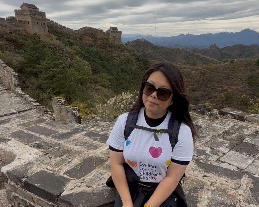 Julie is smiling while wearing sunglasses and a white Evelina London Children’s Charity T-shirt, sitting on a section of the Great Wall of China. The wall stretches behind her through greenery.