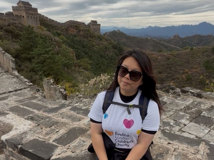 Julie is smiling while wearing sunglasses and a white Evelina London Children’s Charity T-shirt, sitting on a section of the Great Wall of China. The wall stretches behind her through greenery.