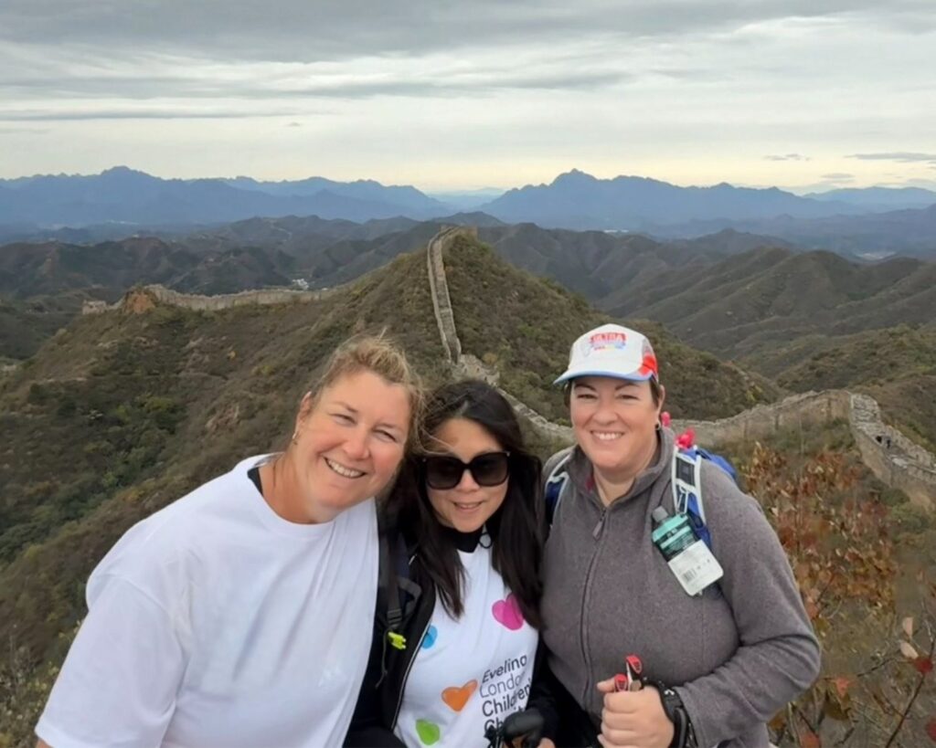 Three women are pictured in front of The Great Wall of China, in the middle is Julie wearing sunglasses and white t-shirt that reads Evelina Children