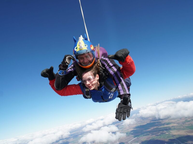 Evelina London Charity supporter skydiving, they're in a red jumsuit with black gloves on and a blue vest. They're with a skydiving instructor who is wearing a helmet.