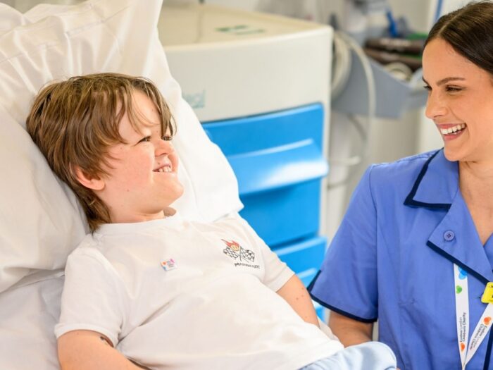 Oliver in a white t shirt appears to be smiling, laid in hospital bed besides him is nurse wearing blue uniform with a yellow name badge that reads 