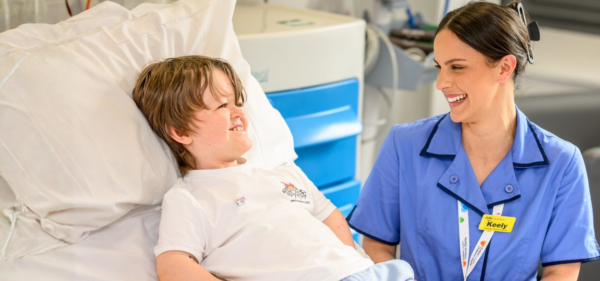 Oliver in a white t shirt appears to be smiling, laid in hospital bed besides him is nurse wearing blue uniform with a yellow name badge that reads 'Keely'.