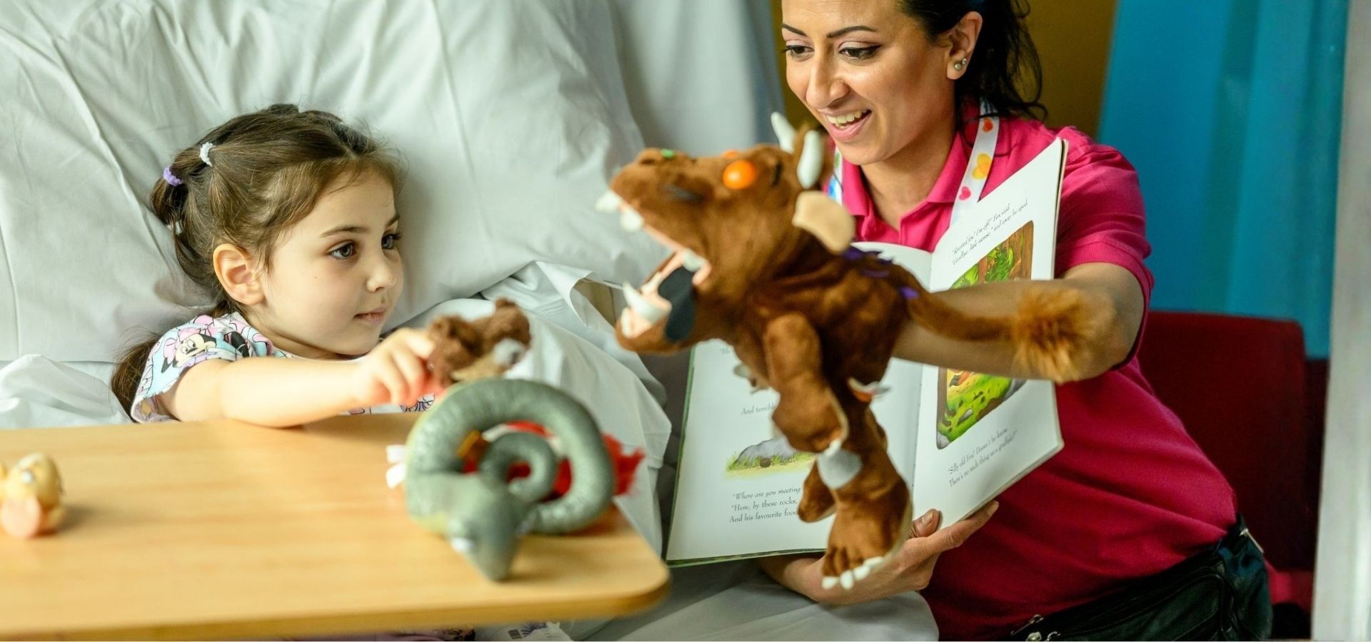 Olilia in Evelina London Children's Hospital bed, beside her is play specialist Shirin with a story book and holding a gruffalo hand puppet reading to Olilia the gurffalo story.
