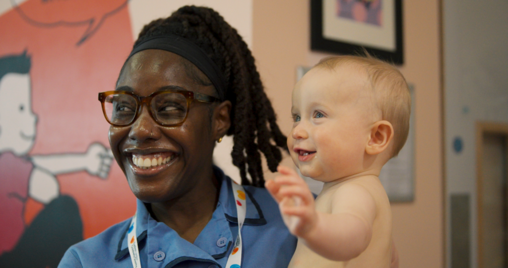 Susanah wearing glasses, blue uniform and white lanyard and appears to be smiling, carry in her arms Baby Ash