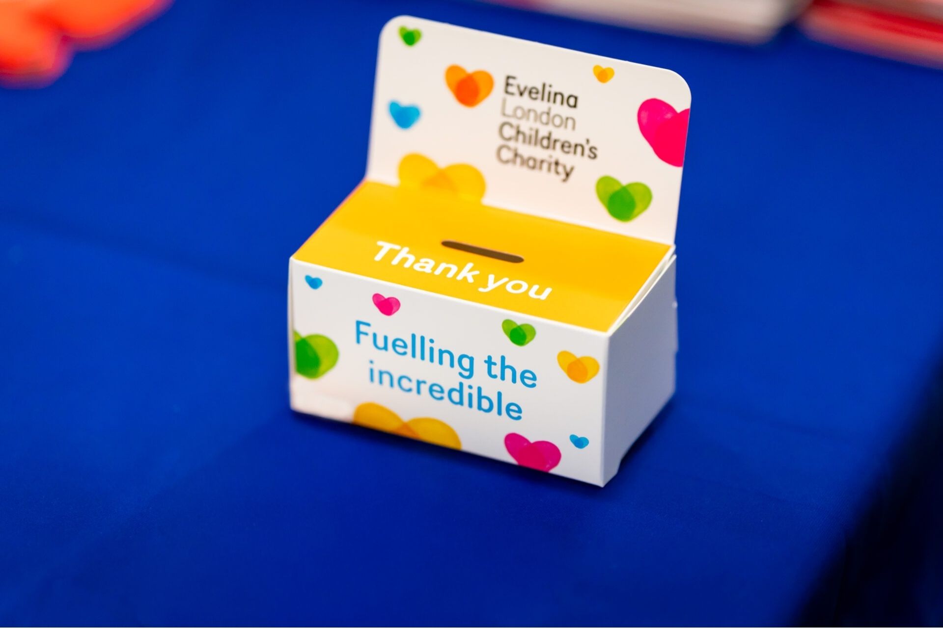 Evelina London Children's Charity donations collection box, the top of the box has a slot and reads 'Thank you' in white text on a yellow background. On the front of the box there is heart graphic motifs in various colours.
